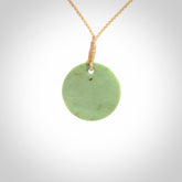 This piece is a small oval round, disc pendant carved in New Zealand Jade. It is suspended on a beige coloured braided cord that is length adjustable.